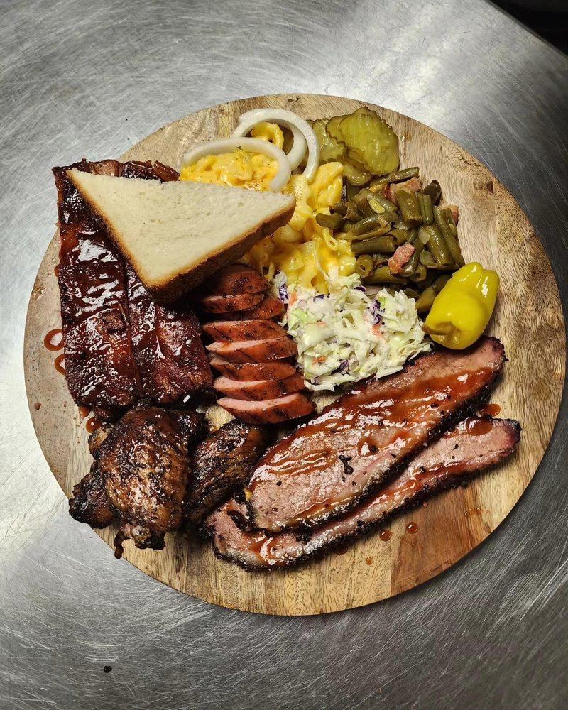 Barbecue platter on a wooden board: ribs, sliced brisket, sausage, coleslaw, mac and cheese, pickles and white bread